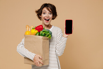 Young amazed woman in casual clothes hold paper bag with vegetables hold in hand use mobile cell...