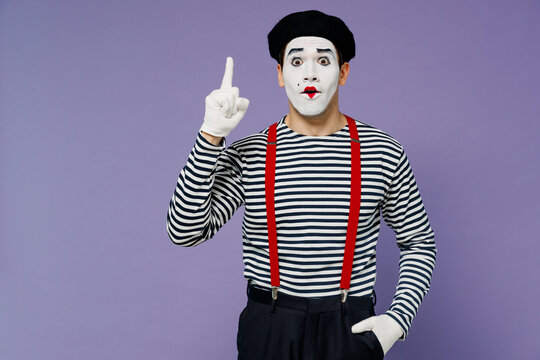 Surprised fun ecstatic young mime man with white face mask wears striped shirt beret pointing up on workspace area copy space mock up isolated on plain pastel light violet background studio portrait.