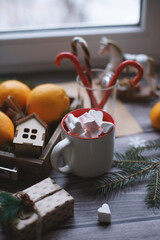 A mug with marshmallows, caramel canes, toys, oranges in a box stand on a light background near the window.