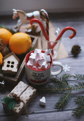 A mug with marshmallows, caramel canes, toys, oranges in a box stand on a light background near the window.