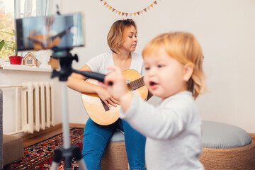 A young mother playing the guitar and records a live video broadcast on a smartphone. A child in a blur in the foreground. The concept of video streaming and online learning