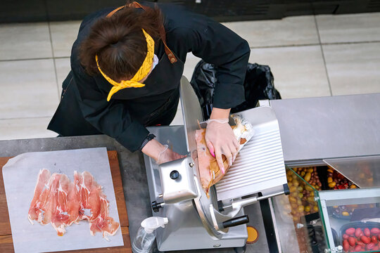 Butcher slicing prosciutto on cutting machine, woman cut prosciutto, . View from above on female butcher cutting meat at butcher counter. Selective focus