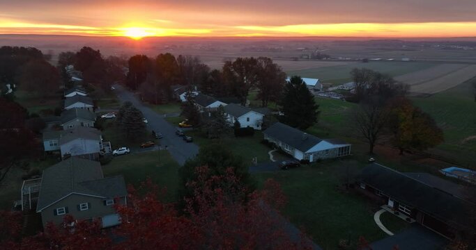 Aerial reveal of houses along quiet street during rural sunrise. Colorful autumn fall foliage and homes in USA American community.