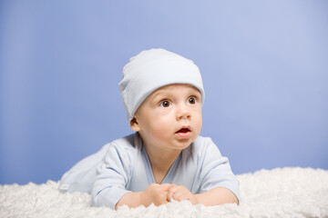 A cute baby in blue clothes and a blue hat lies on a white bedspread. Portrait of a six month old boy, studio shooting