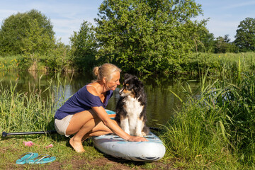 A tanned woman in a t-shirt and shorts. She prepares the sup board for sailing. Her german shepherd dog is sitting on the paddle board. animal themes, sports and leisure. Waterfront, reed, river, 