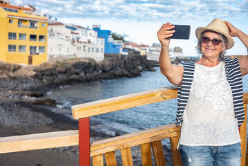Fototapeta na wymiar Smiling senior woman using smart phone por photography in outdoors at sea leaning at a wooden rail. Blue sky and horizon over water