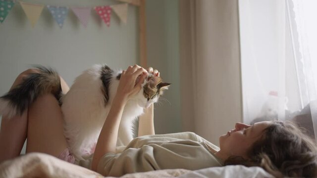 Young brunette curly-haired woman lies on bed covered with white woolen blanket and caresses white fluffy cat holding pet gently