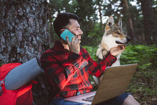 Man with laptop talking on mobile phone while sitting by dog in forest