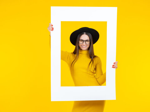 Happy woman holds a white frame in front of her face. Smiling girl. Girl in a yellow dress and a black hat. The lady poses as in the picture. A bright studio shot of a long-haired positive model.