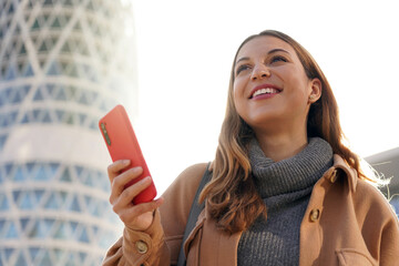 Young confident business woman looking up to the bright future. Excited beautiful lady holding telephone in her hand with modern skyscraper on background.