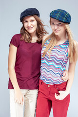 Youth Lifestyle. Two Caucasian Girlfriends Embracing Together While Wearing Hats Over White Background.
