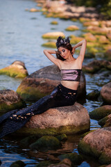 Asian Mermaid Posing At Ocean Shore on Rocks While Wearing Seashell Decorated Crown and Black Shiny Tail On Seductive Body Covered With Strasses