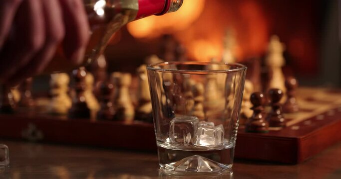 Glass whiskey, scotch, bourbon or cognac stands on a table against background of chess and a fireplace with a flame. Ice near a glass. Alcoholic drink concept. Close up. 4K