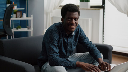 Portrait of black man in living room typing on laptop, looking up at the camera and smiling. African man working from home, computer user in flat apartment using social media freelancer in slow motion