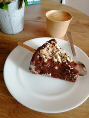 
On a white plate is a piece of chocolate cake sprinkled with nuts. There is a paper cup with aromatic coffee nearby