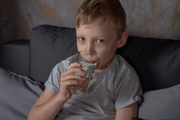 A caucasian boy drinks water from a glass while sitting on a gray sofa