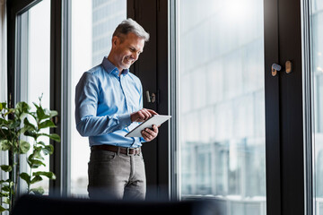 Smiling mature businessman working on digital tablet by window at work place