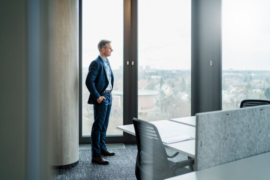 Mature businessman with hands in pockets looking through window in office