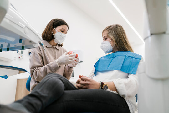 Female orthodontist wearing face mask explaining artificial teeth model to patient at office