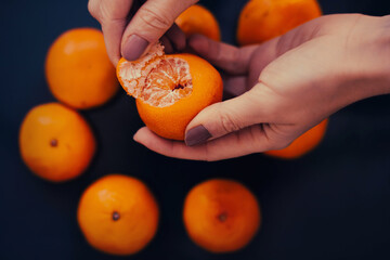 A woman holds a ripe delicious tangerine in her hands and peels it from the peel, against the background of a table on which there are several more tangerines. Citrus harvest. Fruits for Christmas.