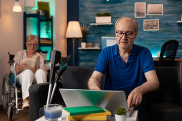 Senior retired man using laptop on table in living room. Aged person with modern device and crutches on couch while disabled old woman sitting in wheelchair in background at home.