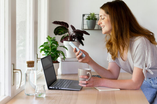 Smiling woman using smart phone over laptop while leaning on desk at home office