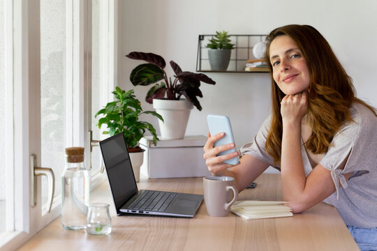 Beautiful woman with hand on chin holding smart phone at desk in home office