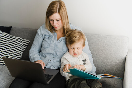 Mother using laptop while sitting son reading picture book on sofa at home