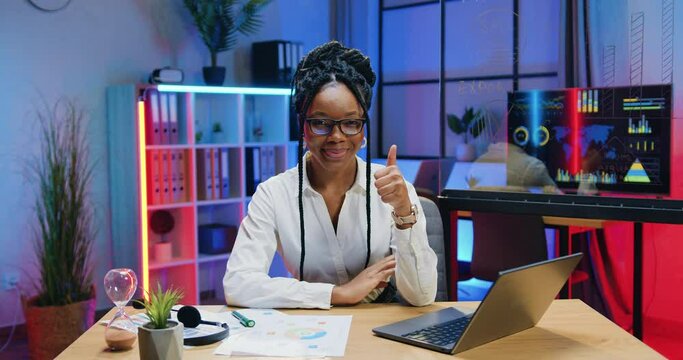 Human portrait of attractive smiling happy successful high-skilled stylish african american businesswoman in glasses which gesturing thumbs up into camera in evening office