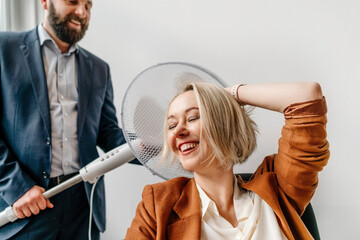 Cheerful businesswoman with eyes closed sitting by male colleague holding electric fan in office