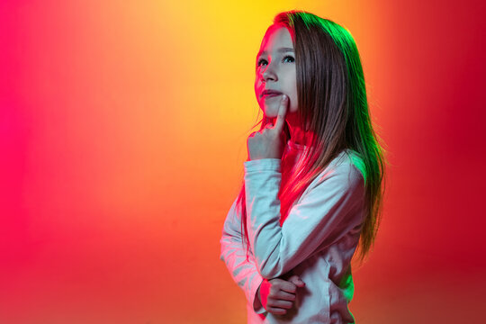 Half-length portrait of little thoughtful girl dreaming isolated on mangenta yellow studio background in neon light, filter.