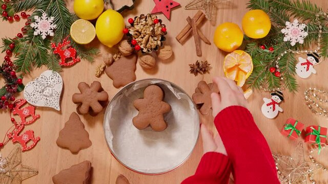 Girl's hands laying Christmas gingerbread cookies in a festive box on a table decorated with Christmas decorations and festive food, top view