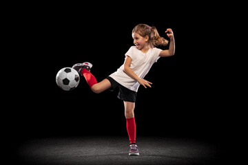 Full-length portrait of little playful girl kicking ball with feet isolated over black background