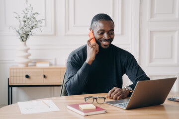 Successful afro american businessman having pleasant telephone conversation while working on laptop