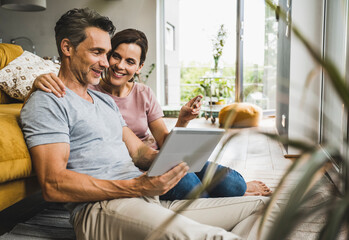 Woman holding credit card while sitting with man using digital tablet at home