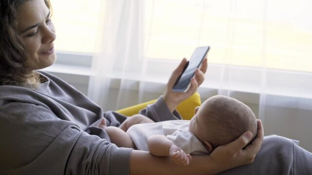 Mother relaxing at home. Side view woman smiling and take photos with smartphone of funny baby girl sleeping on knees while sitting in large armchair against window