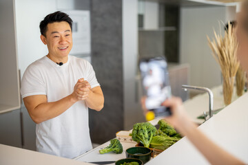 Woman taking video on smartphone of asian man cooking vegetarian salad on kitchen. Concept of video blogging. Idea of healthy and diet eating. Adult male blogger with grapes looking at mobile phone