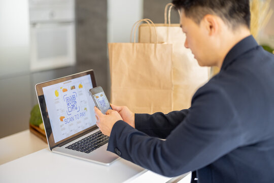 Man using smartphone for payment in online store on laptop on kitchen at home. Concept of online shopping and e-commerce. Idea of healthy and vegetarian eating. Person sit at table with shopping bags