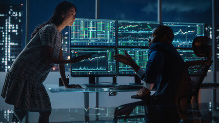 Financial Analyst Talking to Investment Banker in the Evening at Work. Chatting About Real-Time Stock Chart Data on Multi-Monitor Workstation. Businesspeople Have a Meeting in Broker Agency Office.