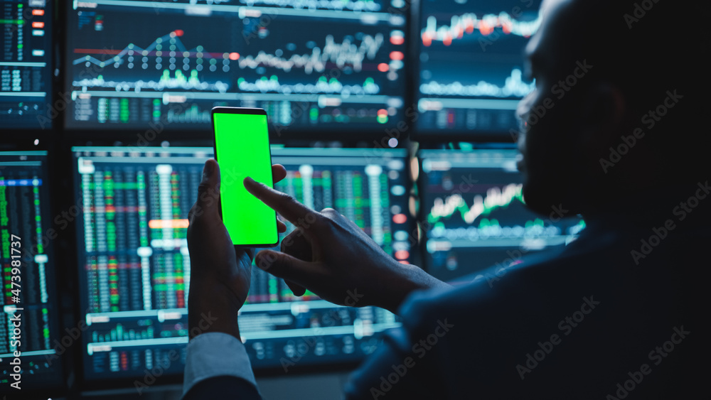 Poster Financial Analyst Using Smartphone with Green Screen Chroma Key Mock Up Template and Working on Multi-Monitor Workstation with Stocks Charts. Businessman Works in Investment Bank in the Evening.