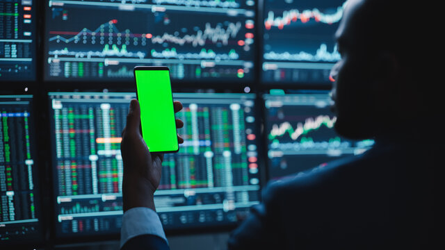 Financial Analyst Using Smartphone with Green Screen Chroma Key Mock Up Template and Working on Multi-Monitor Workstation with Stocks Charts. Businessman Works in Investment Bank in the Evening.