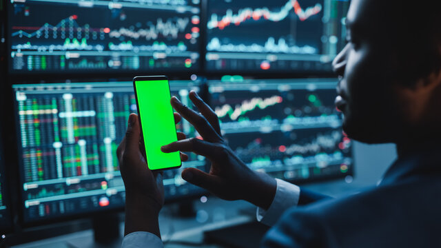 Financial Analyst Using Smartphone with Green Screen Chroma Key Mock Up Template and Working on Multi-Monitor Workstation with Stocks Charts. Businessman Works in Investment Bank in the Evening.