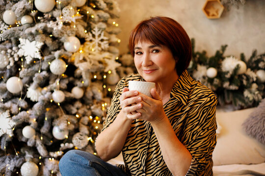 Middle aged beautiful mature woman drinking tea or coffee sitting on sofa over christmas tree blurred background. Christmas and New Year lifestyle concept.