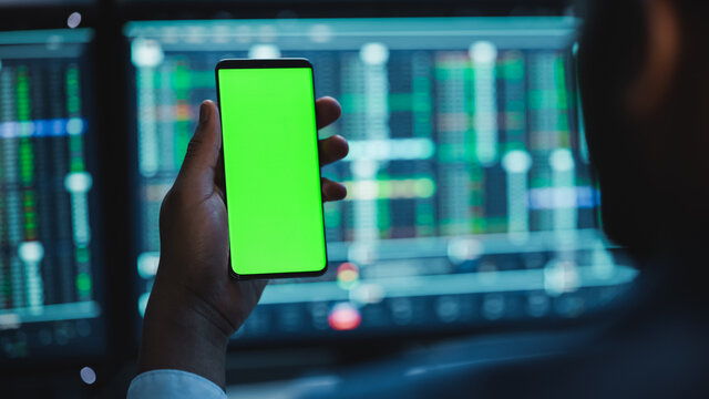 Financial Analyst Using Smartphone with Green Screen Chroma Key Mock Up Template and Working on Multi-Monitor Workstation with Stocks Charts. Businessman Works in Investment Bank in the Evening. - Powered by Adobe