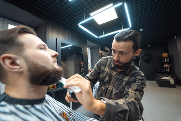 Barber works with a beard clipper. Hipster client getting haircut. - obrazy, fototapety, plakaty