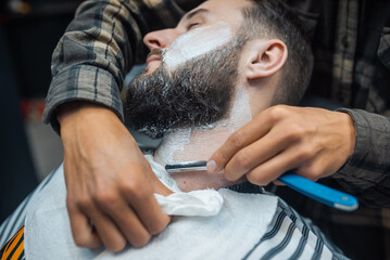 Young bearded man getting shaved by hairdresser at barbershop
