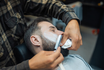 Young bearded man getting shaved by hairdresser at barbershop