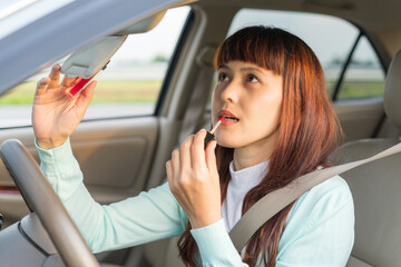 Fototapeta na wymiar Young woman looking in rear view mirror and making up her lips while sitting behind the wheel of her car. Female painting lips doing applying make up while driving the car. Concept of danger driving.