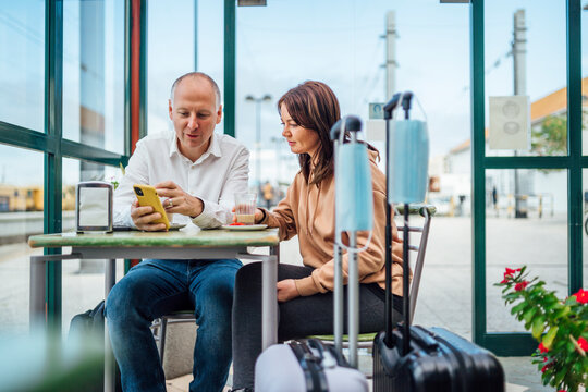 A couple of travelers drinking coffee and checking something on the phone. The luggage with masks in the foreground.