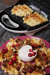 Waffles with sour cream and cranberries. Lie on a plate. Next to it is a waffle maker with waffles. On black pine boards.